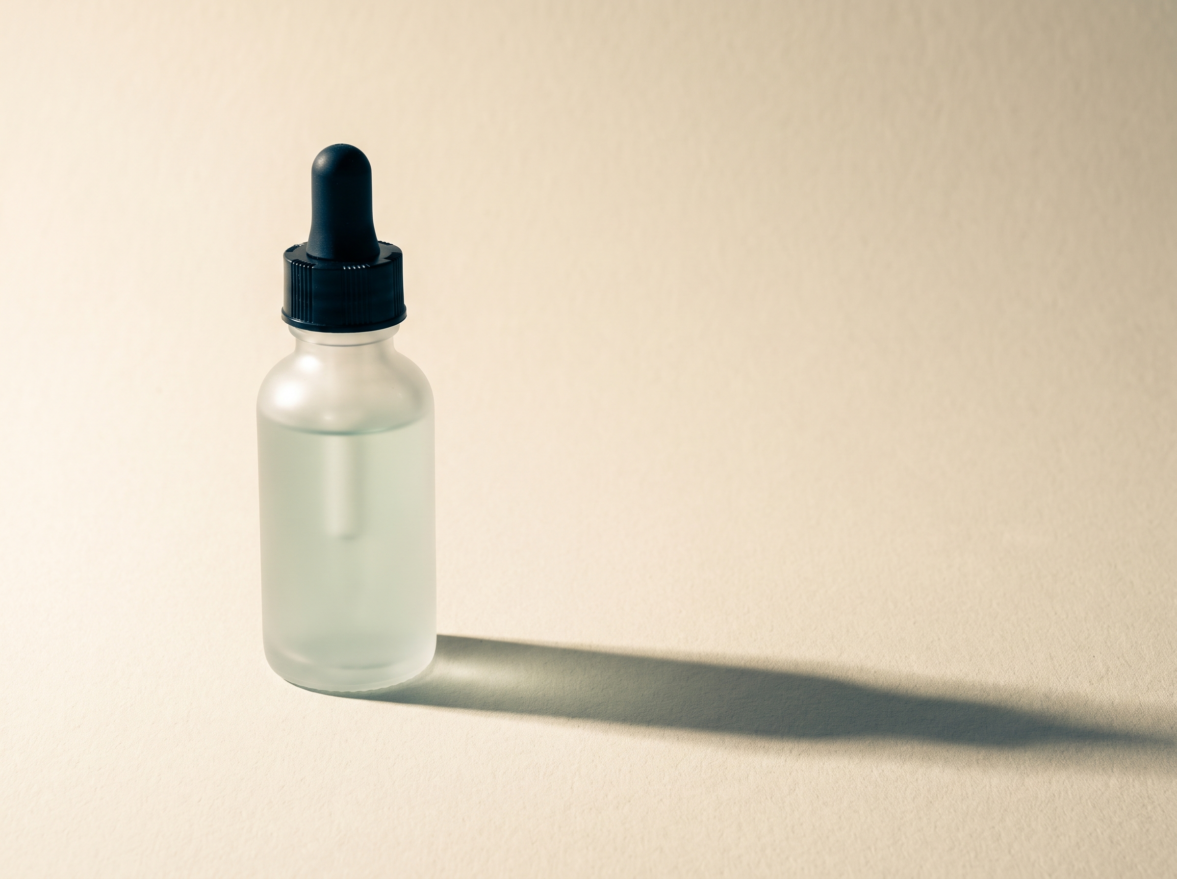 A frosted glass serum bottle with a black dropper cap on cream paper in cold side light